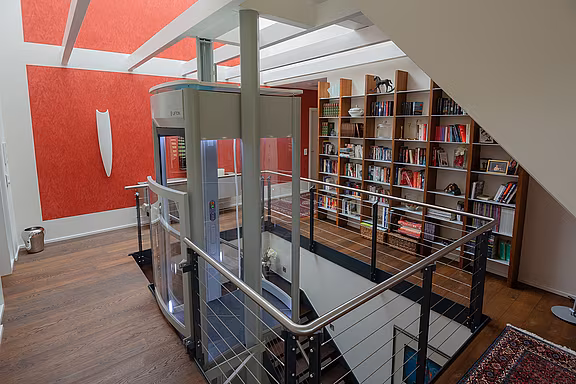 Ein moderner Homelift steht still auf einer Galerieebene, umgeben von einem Bücherregal und einer roten Wand, im Kontext eines stilvollen, offenen Wohnraumes.