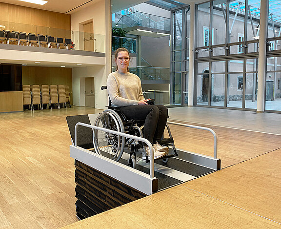 Eine Frau im Rollstuhl fährt auf eine Plattformliftrampe im Innenraum zu, nahe einer Treppe. Im Hintergrund stehen Spinde und ein Laufband.
