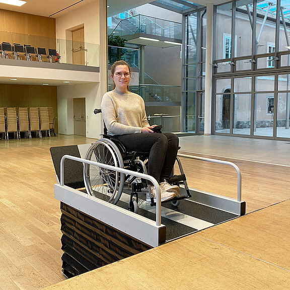 Eine Frau im Rollstuhl fährt auf eine Plattformliftrampe im Innenraum zu, nahe einer Treppe. Im Hintergrund stehen Spinde und ein Laufband.