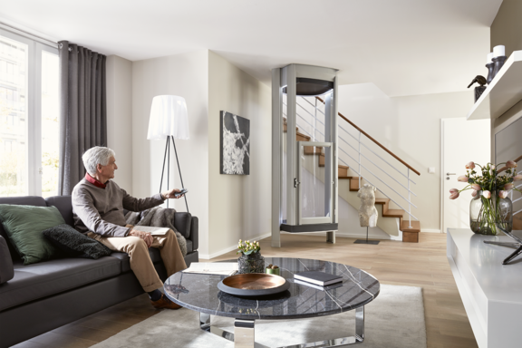 Ein älterer Mann sitzt auf einem Sofa und bedient per Fernbedienung einen modernen Homelift im Wohnzimmer neben einer Treppe; der Homelift steht im ruhenden Zustand.