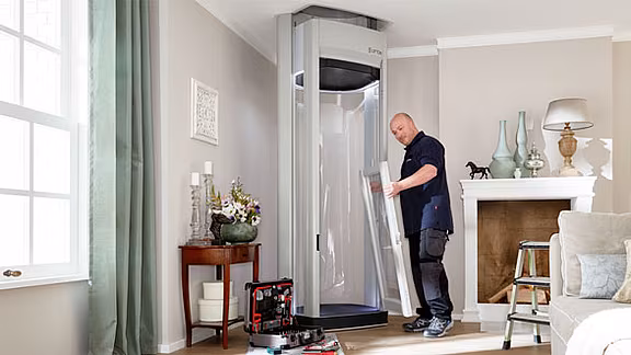 Ein Techniker baut einen Homelift im Wohnzimmer eines Hauses zusammen; Werkzeugkoffer und Bauteile liegen bereit, im Hintergrund Kamin, Dekoration und Tageslicht durch das Fenster.