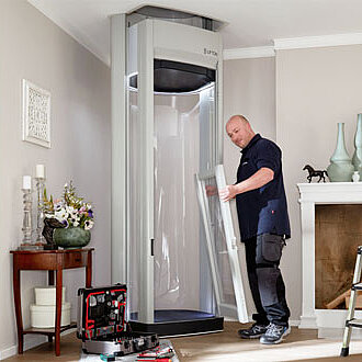 Ein Techniker baut einen Homelift im Wohnzimmer eines Hauses zusammen; Werkzeugkoffer und Bauteile liegen bereit, im Hintergrund Kamin, Dekoration und Tageslicht durch das Fenster.