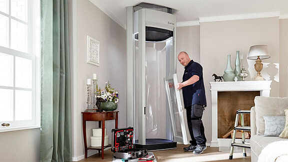 Ein Techniker baut einen Homelift im Wohnzimmer eines Hauses zusammen; Werkzeugkoffer und Bauteile liegen bereit, im Hintergrund Kamin, Dekoration und Tageslicht durch das Fenster.