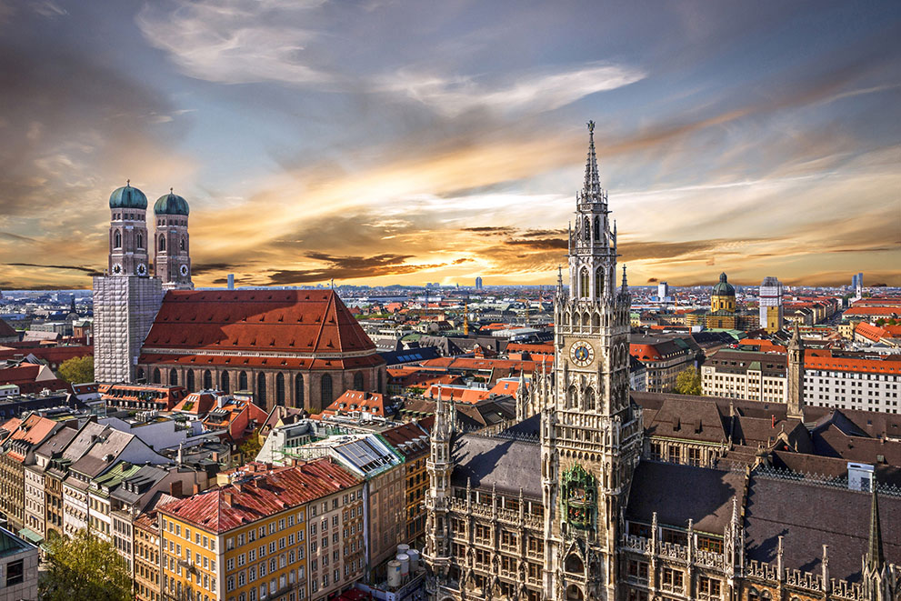 Das Neue Rathaus dominiert das Stadtbild von München im Sonnenuntergang, während die Frauenkirche links im Hintergrund sichtbar ist; zahlreiche Dächer und Gebäude umgeben die Szene.