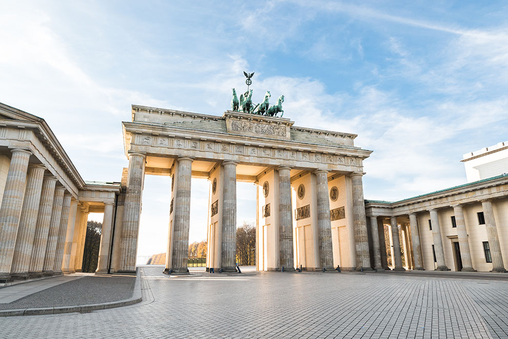 Das Brandenburger Tor steht ruhig und imposant auf einem großen, leeren Platz in Berlin, mit blauem Himmel im Hintergrund.