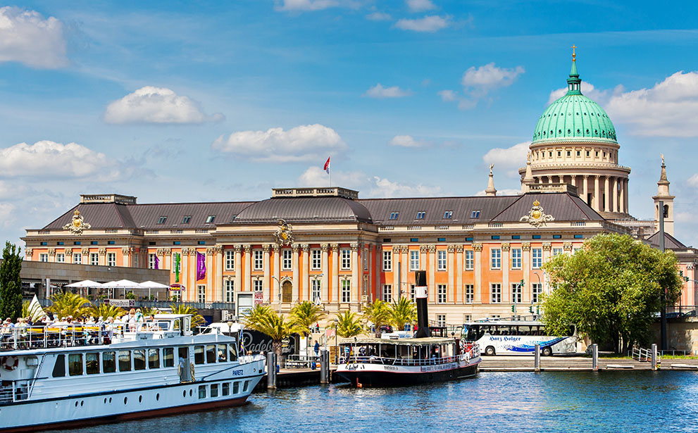 Das Bild zeigt das Alte Rathaus in Potsdam mit markanter Kuppel; im Vordergrund fahren Ausflugsschiffe auf einem Fluss, Passagiere genießen die sonnige Atmosphäre am Wasser.
