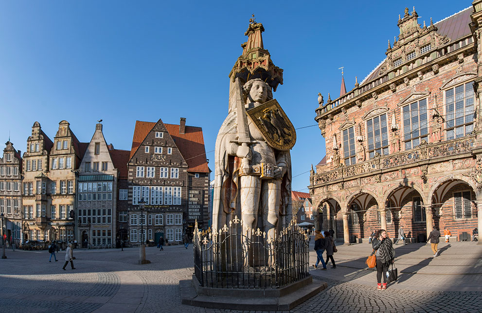 Eine große Rolandstatue steht auf einem Platz in Bremen, mehrere Menschen gehen vorbei, im Hintergrund das historische Rathaus Bremen mit verzierten Fassaden und alten Giebelhäusern