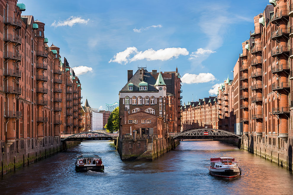 Zwei Boote fahren auf einem Kanal zwischen roten Backsteingebäuden der Speicherstadt Hamburg; zentral steht das bekannte Wasserschloss. Im Hintergrund sind Brücken und die Elbphilharmonie sichtbar.