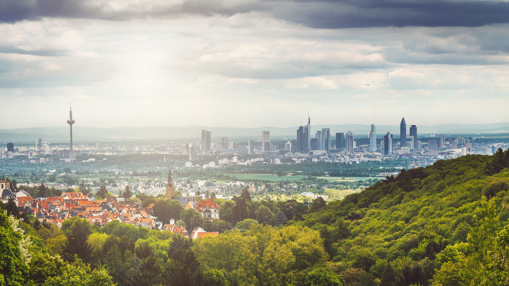 Die Skyline von Frankfurt am Main mit zahlreichen Hochhäusern, darunter das markante Commerzbank Tower, ist von einem grünen, bewaldeten Vordergrund und Vororten umgeben