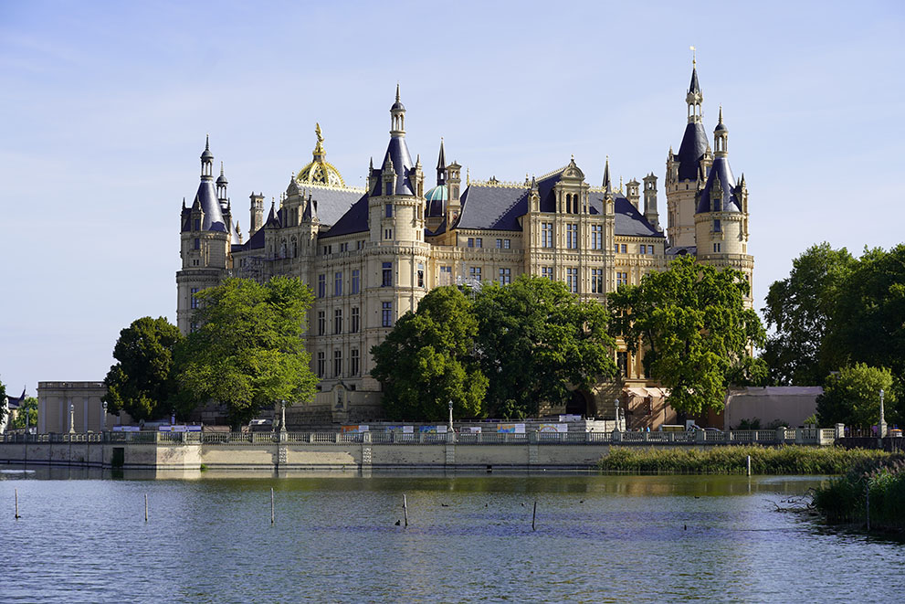 Das Bild zeigt das Schweriner Schloss in der Stadt Schwerin, das majestätisch am Ufer eines Sees steht, umgeben von Bäumen und Wasser, im Tageslicht