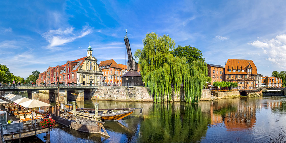 Ein historischer Kran und Fachwerkhäuser stehen am Fluss in Lüneburg; Menschen spazieren auf einer Brücke, Boote liegen am Ufer, links sind Außengastronomie und Sonnenschirme zu sehen