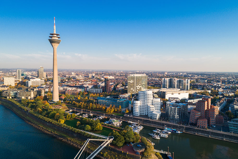 Der Rheinturm steht prominent in Düsseldorf, ragt am Flussufer empor, umgeben von modernen Gebäuden und dem Hafen