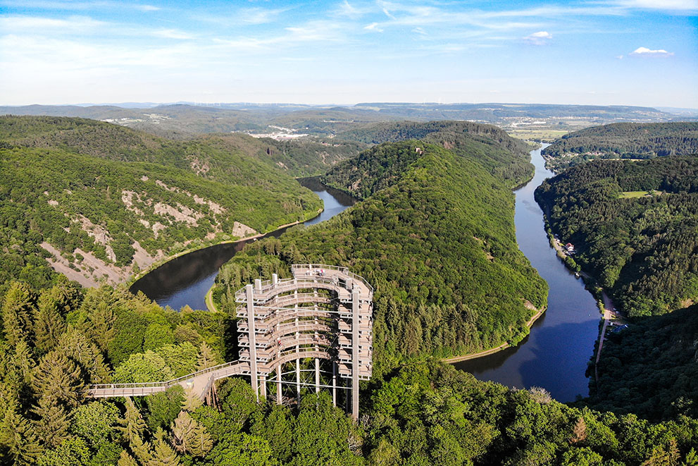 Ein hölzerner Aussichtsturm (Baumwipfelpfad Saarschleife) steht im Vordergrund, Menschen gehen darauf, umgeben von dichten Wäldern und einem Fluss, der eine markante Schleife bildet