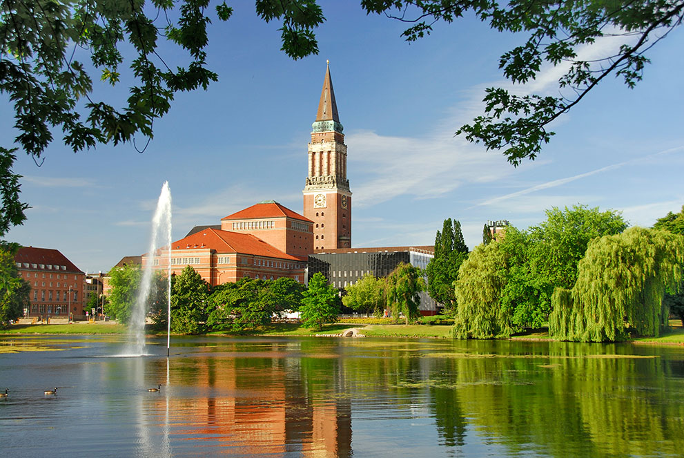 Das Kieler Rathaus steht im Zentrum von Kiel, umgeben von Bäumen; ein Brunnen sprüht Wasser in einen ruhigen See im Vordergrund, Gänse schwimmen darauf