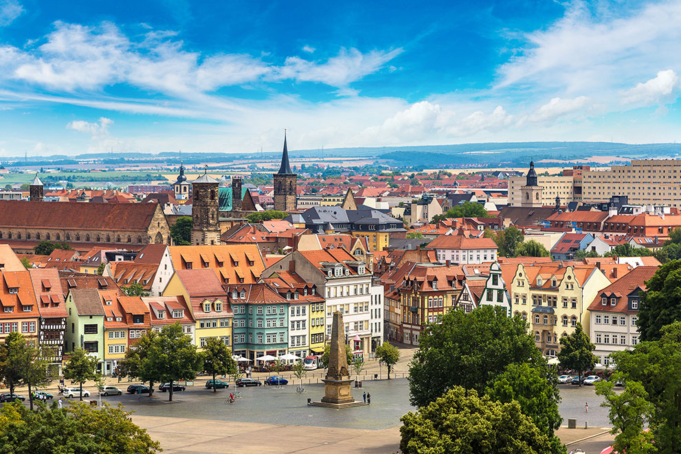 Bunte Fachwerkhäuser stehen am Erfurter Domplatz in Erfurt. Menschen sitzen in Straßencafés, im Zentrum steht der Obelisk. Im Hintergrund ist der Dom St. Marien erkennbar.