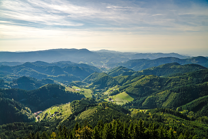 Ein grünes, bewaldetes Mittelgebirge erstreckt sich unter blauem Himmel in Baden-Baden; vereinzelte Höfe und Wiesen liegen in den Tälern