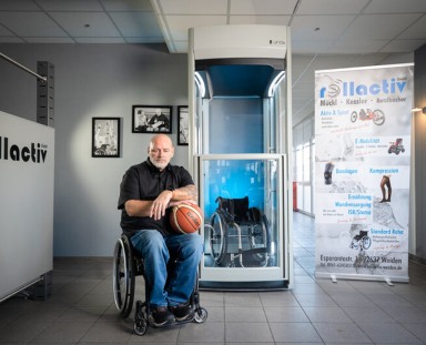 Mann sitzt im Rollstuhl mit einem Basketball in der Hand vor einem Homelift