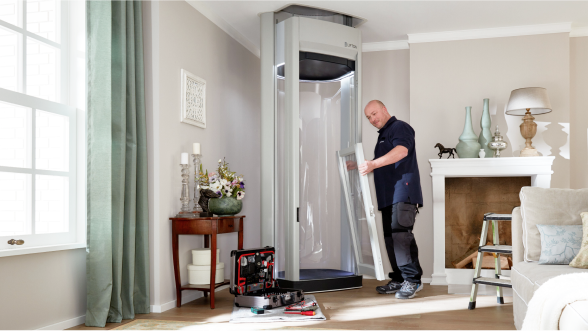 Ein Techniker baut einen Homelift im Wohnzimmer eines Hauses zusammen; Werkzeugkoffer und Bauteile liegen bereit, im Hintergrund Kamin, Dekoration und Tageslicht durch das Fenster.