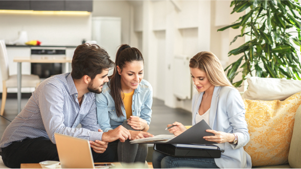 Drei Menschen sitzen an einem Tisch im Wohnzimmer, schauen gemeinsam in einen Ordner und besprechen Unterlagen. Im Hintergrund ist eine Küche sichtbar.