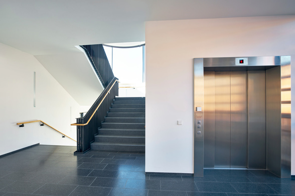 Ein moderner Aufzug mit geschlossener Edelstahltür steht still in einem hellen Treppenhaus, daneben führen Stufen mit schwarzem Geländer nach oben, Tageslicht fällt durch ein Fenster.